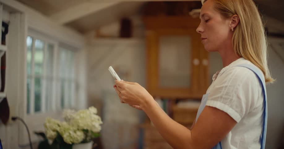 A woman in an apron uses her smartphone to photograph a bouquet of white hydrangeas in her rustic home, shot in slow motion