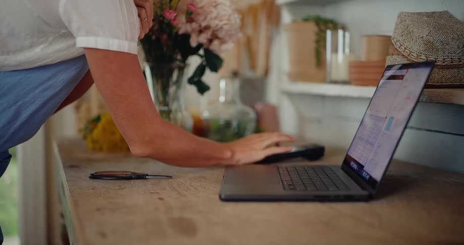 A florist manages her small business on a laptop, standing at a wooden counter surrounded by fresh flowers in her shop