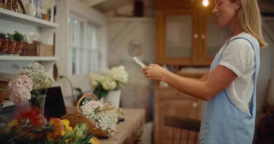 A woman wearing a blue apron uses her smartphone to take photos of flower arrangements in her rustic workshop