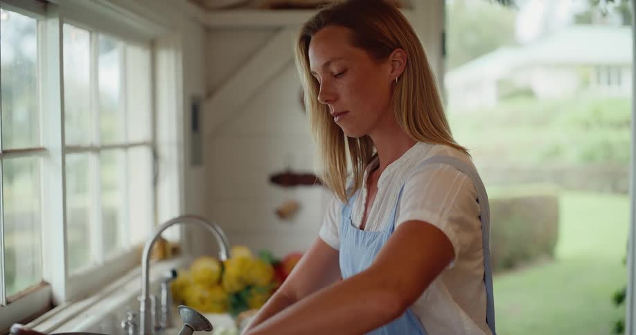 A young woman in a blue apron thoughtfully arranges a bouquet of freshly cut flowers over a sink in a rustic, sunlit room