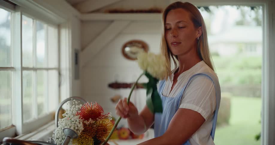 A peaceful woman in a blue apron mindfully arranges a colorful bouquet of flowers in her rustic country kitchen in slow motion