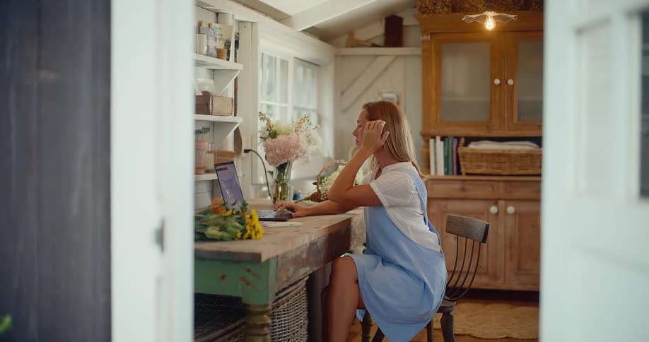 A woman works on her laptop and phone at a rustic desk in her beautiful, sunlit, flower-filled workshop
