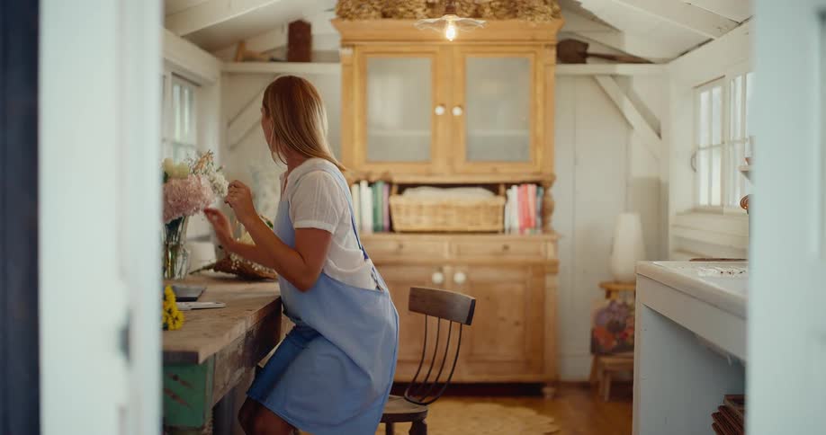 A female florist works on her laptop in her charming workshop before getting up to arrange a fresh bouquet of flowers