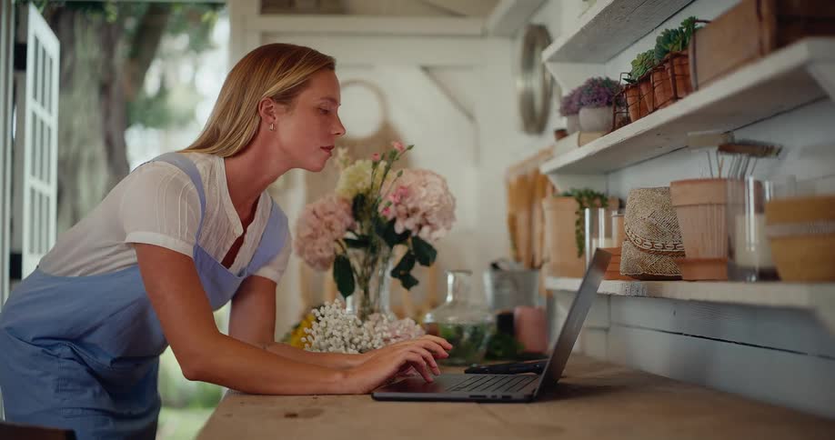 A focused female florist manages her small business online, working on a laptop inside her charming and rustic flower workshop