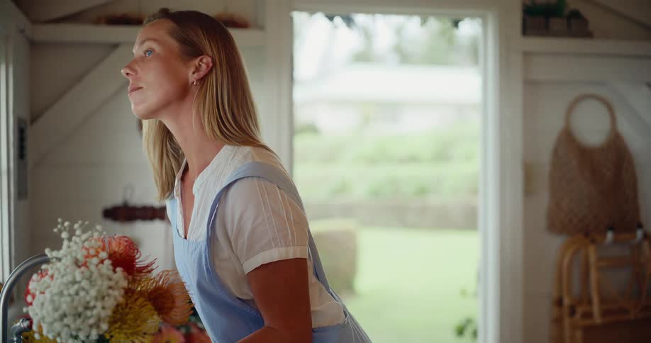 A florist wearing an apron thoughtfully gazes out of a rustic kitchen window while holding a freshly picked bouquet of flowers