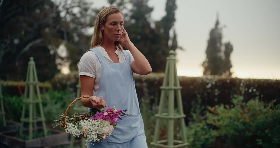 A thoughtful woman wearing an apron holds a basket of fresh flowers while standing peacefully in her beautiful garden at dawn