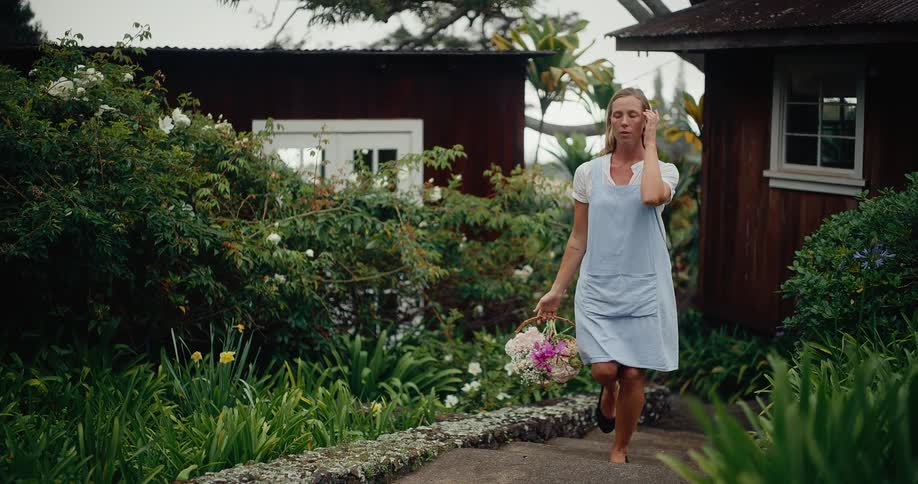 A woman in a blue apron walks thoughtfully through a lush garden path with a basket of fresh-cut flowers in slow motion
