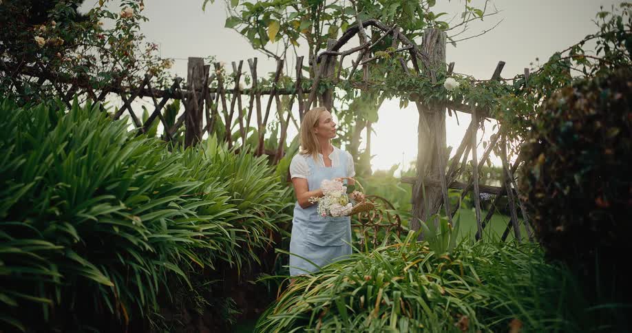 A serene woman in an apron walks through her lush garden at sunset, carrying a basket of freshly picked flowers