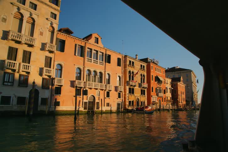 Historic Venetian buildings lining a canal glow in the warm evening sun, viewed from a passing boat