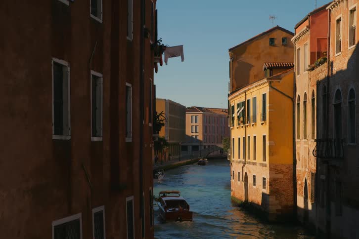 A classic wooden motorboat navigates a sun-drenched canal in Venice, Italy, surrounded by historic, colorful buildings