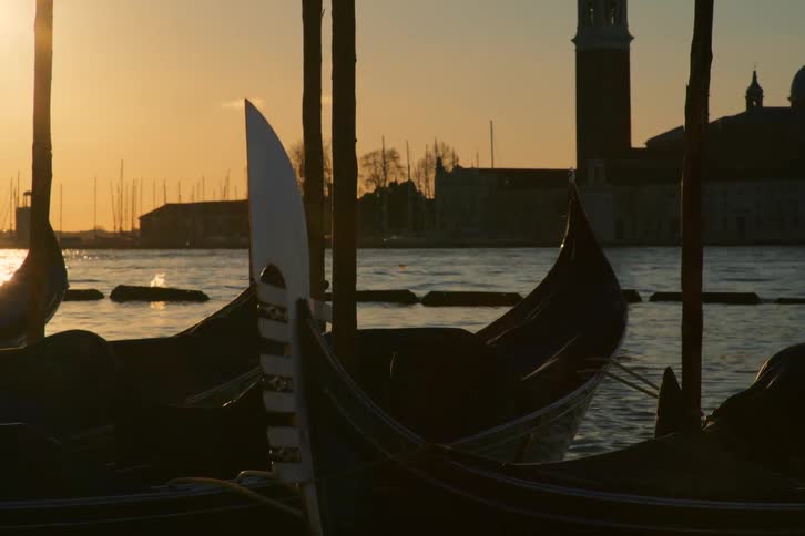 Silhouetted gondolas gently sway in a Venetian canal at sunset, with the Church of San Giorgio Maggiore in the background
