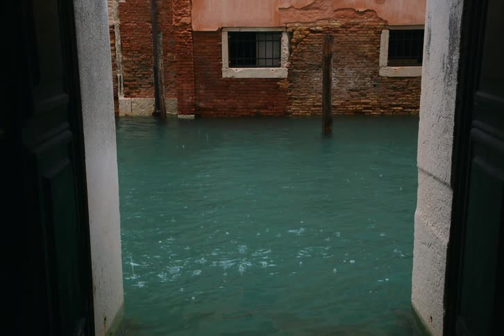 Raindrops gently fall on the flooded teal waters of a canal in Venice, Italy, viewed from a doorway