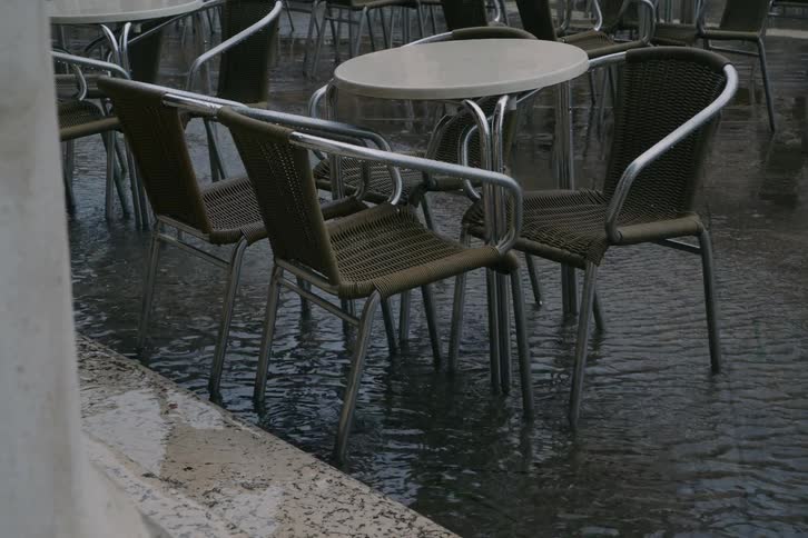 Empty outdoor cafe tables and chairs stand in floodwaters during a heavy rainstorm
