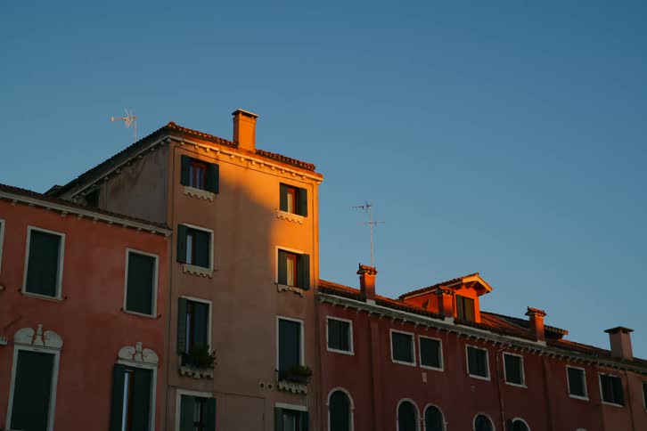 Golden hour sunlight illuminates the rustic rooftops of old Venetian buildings against a clear blue sky