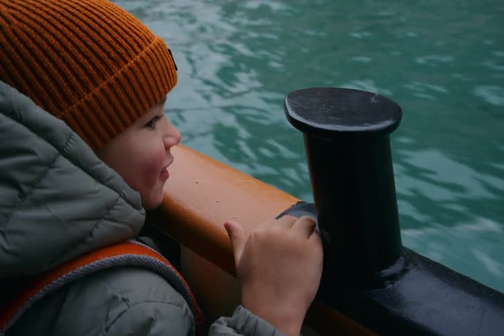 A curious little boy in a warm beanie and life jacket enjoys a boat ride on the lake