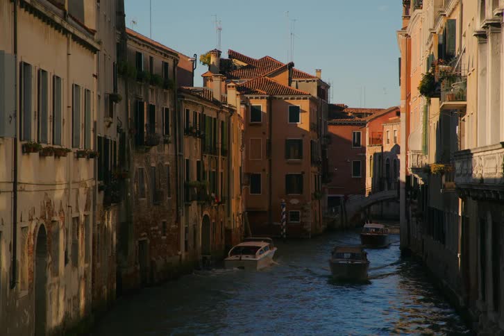 Motorboats cruise down a picturesque Venetian canal flanked by old, sunlit buildings during the golden hour