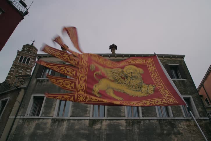 A low-angle view of the iconic Venetian flag with the Lion of Saint Mark waving in the wind against a historic building and overcast sky
