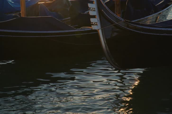 Docked Venetian gondolas gently bob on the water as the golden sunset light reflects on the gentle ripples