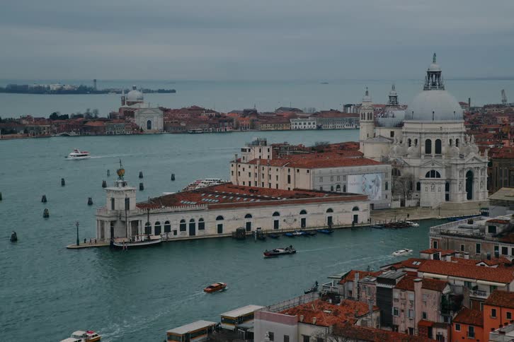 A panoramic high-angle view of the Punta della Dogana and Basilica di Santa Maria della Salute in Venice, with boats navigating the canal on an overcast day