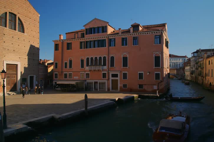 Boats navigate a tranquil canal next to a sunlit square and historic buildings in Venice, Italy