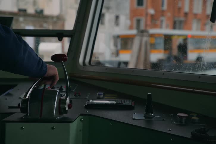 A boat captain's hand operates the throttle and controls in the wheelhouse while navigating a city canal