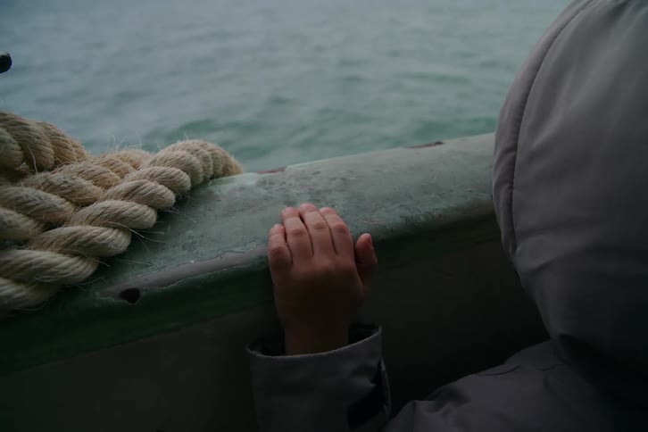 A child's small hand rests on the railing of a boat sailing on a choppy, grey sea on an overcast day