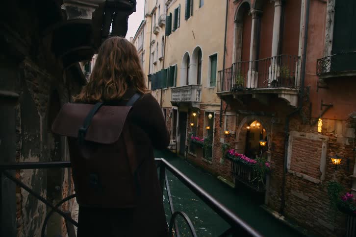 A traveler with long hair and a backpack stands on a balcony overlooking a narrow, moody canal in Venice, Italy