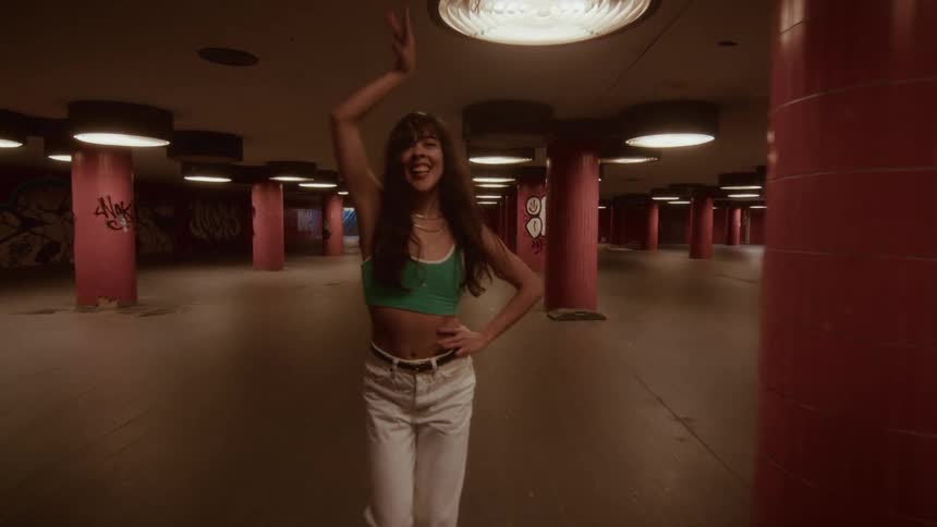 A stylish young woman with long dark hair joyfully dances in slow motion through a gritty, graffiti-covered underground walkway