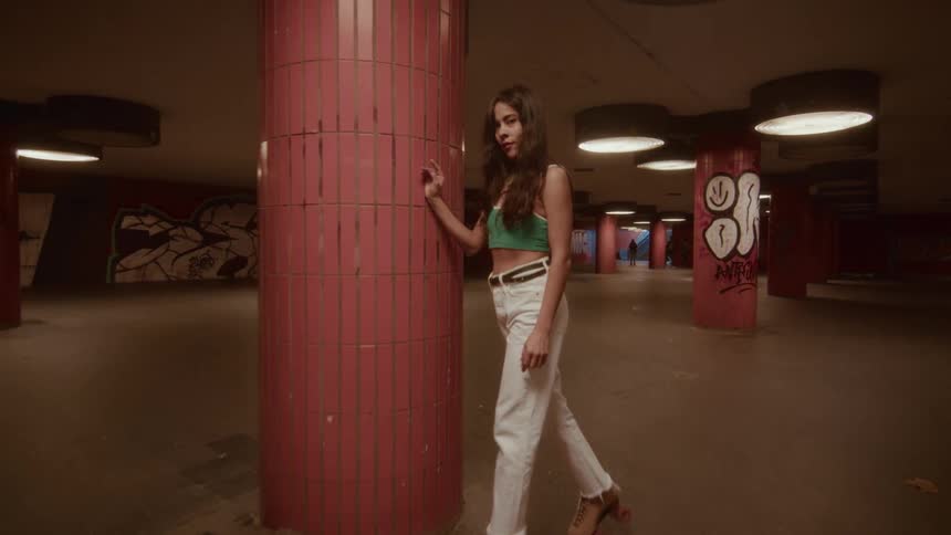 A fashionable young woman on roller skates looks at the camera while leaning on a tiled pillar in a dimly lit, graffiti-filled underpass