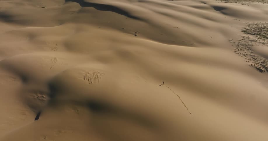 High aerial view of a lone person leaving a trail of footprints as they walk across a vast and desolate sand dune landscape