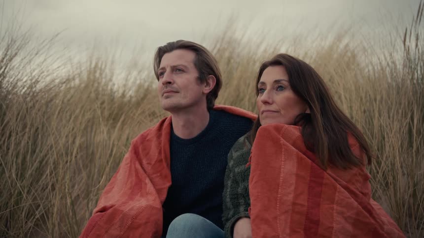 A pensive couple shares a warm blanket while sitting together on a grassy sand dune on a cool day