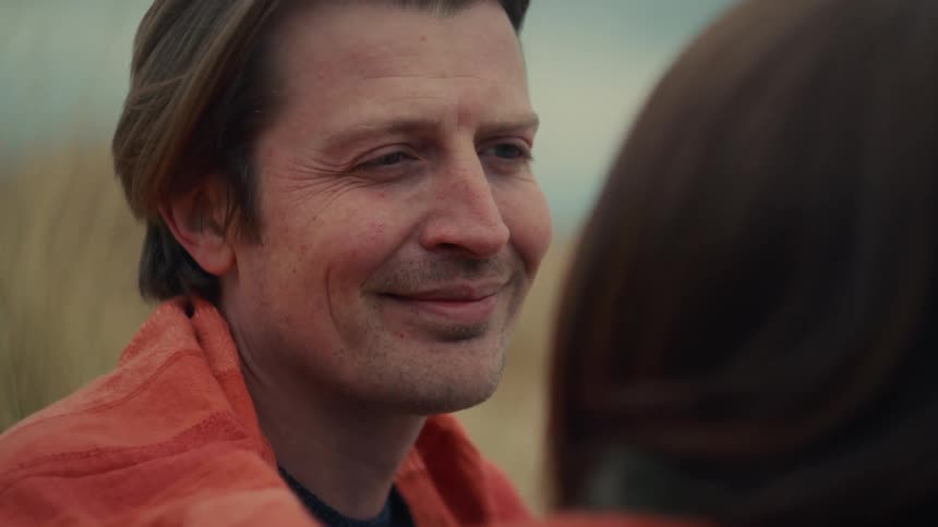 A man wrapped in an orange towel smiles lovingly at his partner during an intimate moment outdoors
