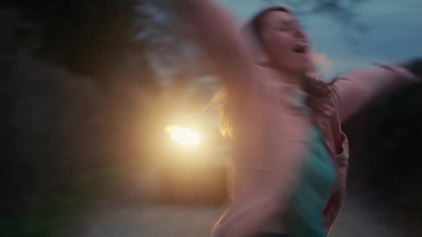 A happy woman spins around with her arms outstretched on a country road at dusk, backlit by the golden glow of headlights in slow motion