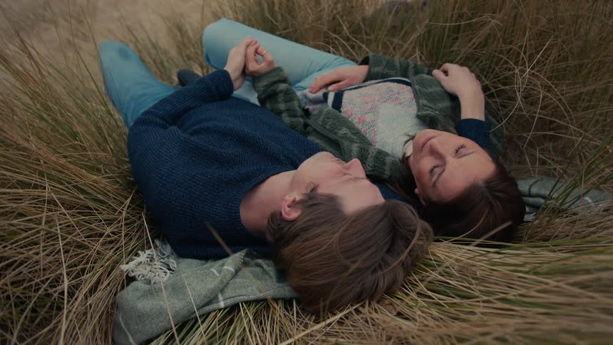 A high-angle view captures a loving couple as they peacefully lay together in tall beach grass, holding hands