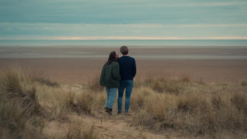 From a grassy dune, a couple peacefully watches the ocean on an overcast day
