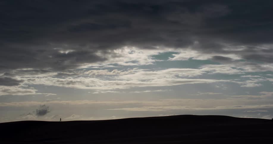 A lone silhouetted figure walks along the horizon of a vast dune landscape under a dramatic and moody sky