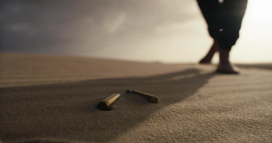 A man walks barefoot across a desert landscape at sunset and kneels to pick up spent bullet casings from the sand in slow motion