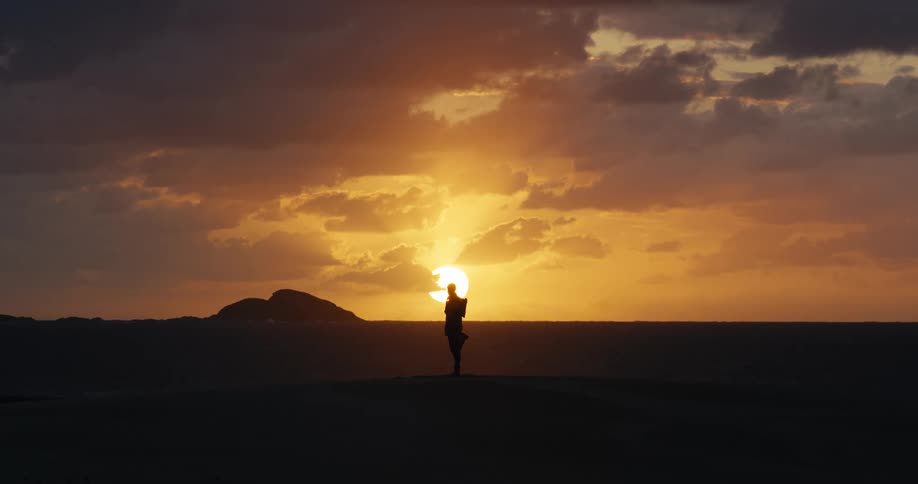 A dramatic wide shot shows the silhouette of a person standing on a dune, appearing to hold the setting sun over the ocean