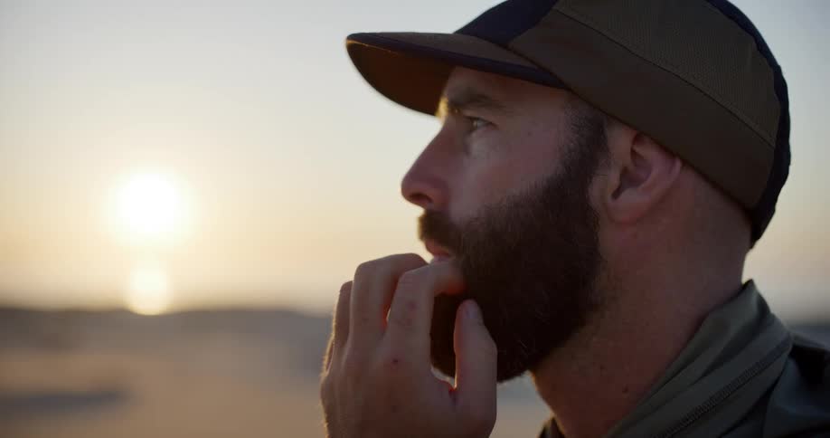 Close up of a pensive bearded man in a hat looking out at the golden sunrise