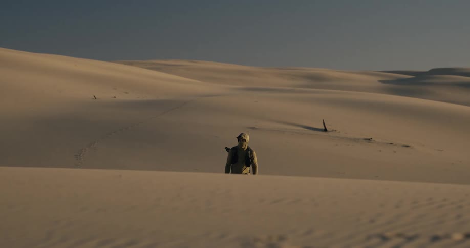 A hooded photographer slowly walks over a sand dune in a vast, empty desert during golden hour