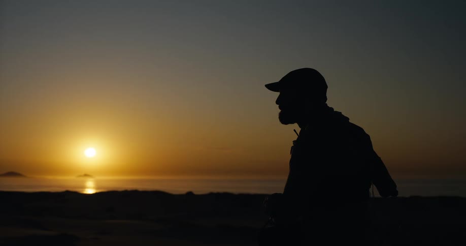 A man in a cap is silhouetted against the rising sun over the ocean as he looks through a pair of binoculars