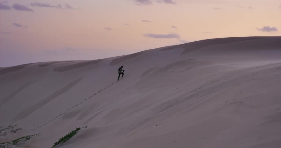 A lone woman with a backpack runs in slow motion up a massive sand dune at sunset, showing determination and endurance
