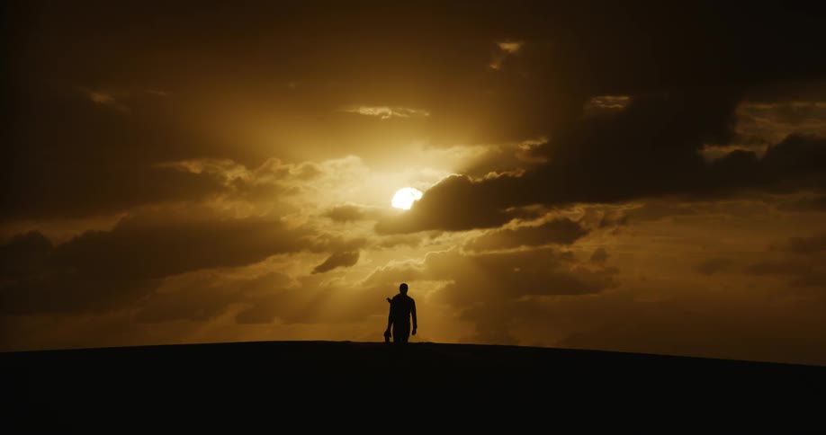 A lone traveler walks over a sand dune, silhouetted against a breathtaking golden sunset