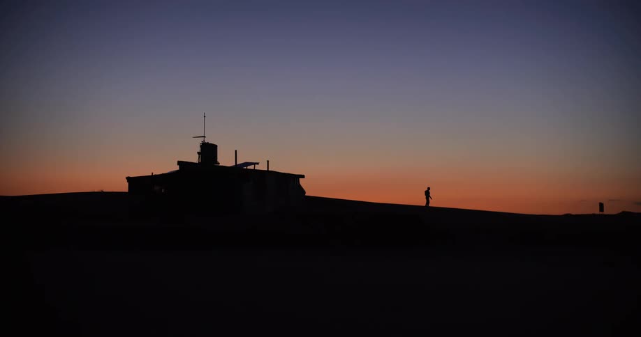 A lone person walks past a remote desert outpost, silhouetted against a beautiful orange and blue sky at twilight