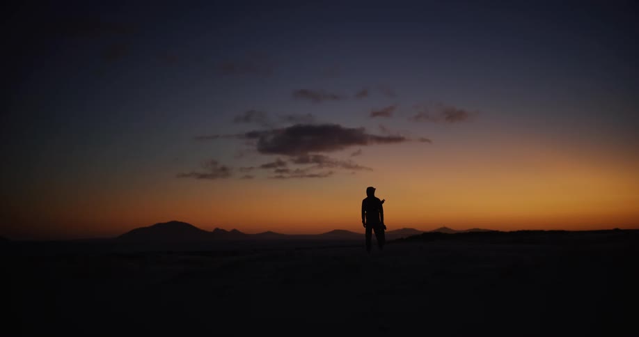 A lone traveler's silhouette walks across a vast desert landscape before stopping to admire the fiery sunset over distant mountains