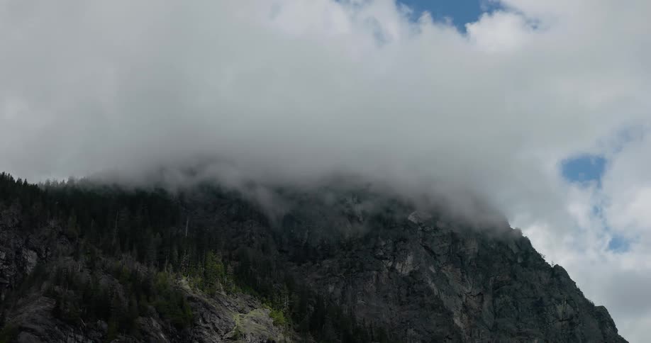 A timelapse of thick, dramatic clouds rolling over the peak of a rugged, tree-covered mountain