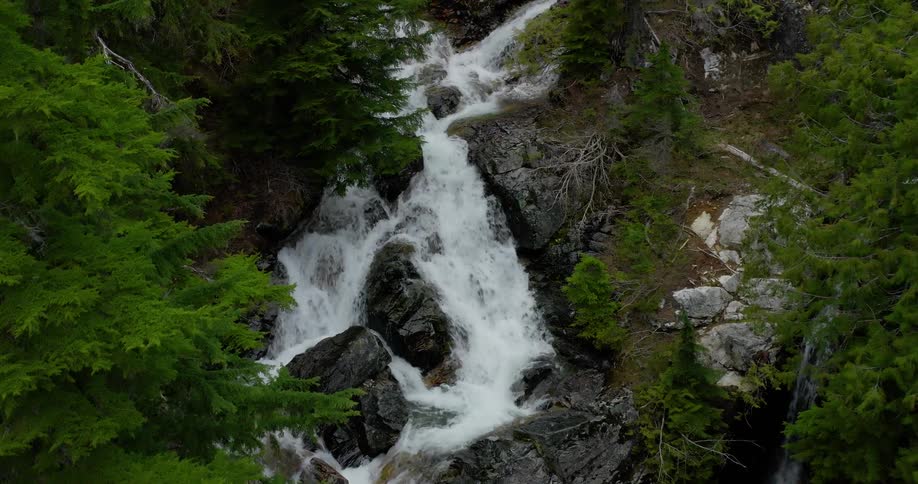 Aerial view pushes in on a beautiful cascading waterfall flowing through a lush green evergreen forest in the mountains