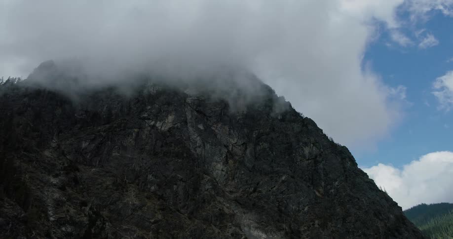 A moody, slow pull-back shot reveals a massive rocky mountain with its peak enveloped in thick clouds