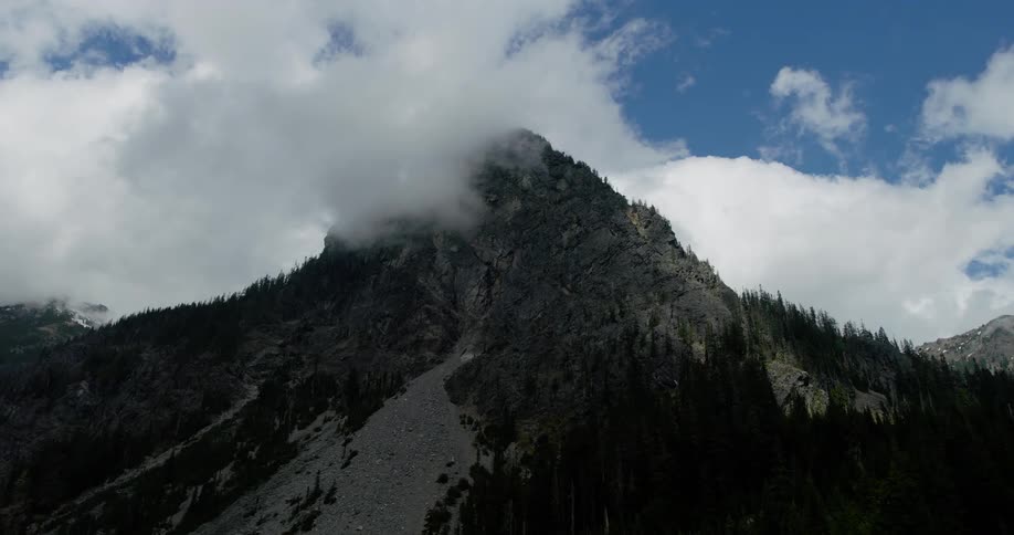 An upward aerial reveal of a rugged mountain peak with a large rockslide, its summit shrouded in dramatic, thick clouds