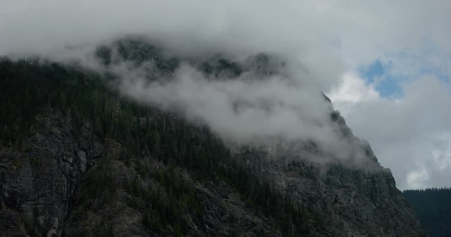 A moody time-lapse shows thick clouds and fog rolling across a rugged, pine-covered mountain